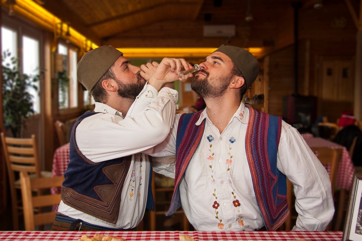 Two men in traditional Serbian outfits drink rakija travelling to Serbia